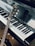 A close-up view of a piano keyboard with a guitar resting against it. The piano keys are clean and polished, reflecting light. The guitar, positioned diagonally, has a wooden body and the brand name is visible on the headstock.