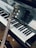 A close-up view of a piano keyboard with a guitar resting against it. The piano keys are clean and polished, reflecting light. The guitar, positioned diagonally, has a wooden body and the brand name is visible on the headstock.
