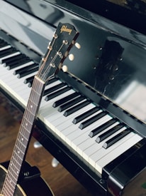 A close-up view of a piano keyboard with a guitar resting against it. The piano keys are clean and polished, reflecting light. The guitar, positioned diagonally, has a wooden body and the brand name is visible on the headstock.