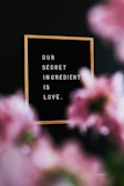 A wooden-framed message board reads 'OUR SECRET INGREDIENT IS LOVE,' with blurred pink flowers in the foreground and a dark background.