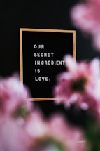 A wooden-framed message board reads 'OUR SECRET INGREDIENT IS LOVE,' with blurred pink flowers in the foreground and a dark background.