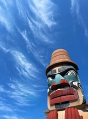 A large, colorful totem pole with intricate carved details stands against a bright blue sky adorned with wispy clouds. The totem features vivid colors such as red, blue, teal, and beige, with expressive facial features.