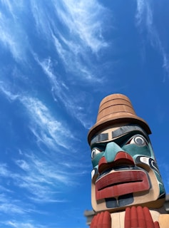 A large, colorful totem pole with intricate carved details stands against a bright blue sky adorned with wispy clouds. The totem features vivid colors such as red, blue, teal, and beige, with expressive facial features.