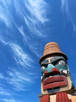 A large, colorful totem pole with intricate carved details stands against a bright blue sky adorned with wispy clouds. The totem features vivid colors such as red, blue, teal, and beige, with expressive facial features.