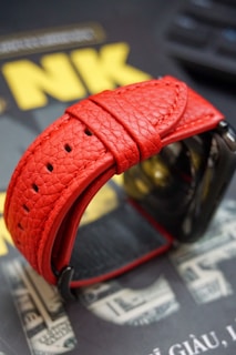 A close-up of a textured red leather watch strap, highlighted against a blurred background that includes text and imagery resembling currency.