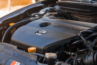 Close-up view of a car engine showing the engine cover with a manufacturer's logo and the text indicating specifications. The surrounding components of the engine are visible, such as wires, hoses, and caps.