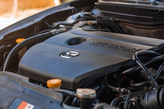 Close-up view of a car engine showing the engine cover with a manufacturer's logo and the text indicating specifications. The surrounding components of the engine are visible, such as wires, hoses, and caps.