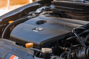 Close-up view of a car engine showing the engine cover with a manufacturer's logo and the text indicating specifications. The surrounding components of the engine are visible, such as wires, hoses, and caps.