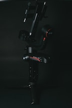 A black, high-tech camera gimbal is set against a dark background. The gimbal features various controls and buttons, with red accents and a small tripod base for stability.