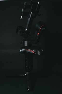 A black, high-tech camera gimbal is set against a dark background. The gimbal features various controls and buttons, with red accents and a small tripod base for stability.