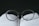 A pair of round-framed eyeglasses is placed on top of an open book. The glasses are positioned in a way that the lenses appear to magnify the text beneath them. The background is dark, creating a strong contrast with the white pages and the clear lenses of the glasses.