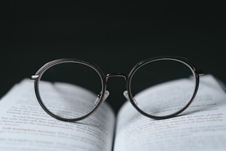 A pair of round-framed eyeglasses is placed on top of an open book. The glasses are positioned in a way that the lenses appear to magnify the text beneath them. The background is dark, creating a strong contrast with the white pages and the clear lenses of the glasses.