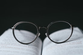 A pair of round-framed eyeglasses is placed on top of an open book. The glasses are positioned in a way that the lenses appear to magnify the text beneath them. The background is dark, creating a strong contrast with the white pages and the clear lenses of the glasses.