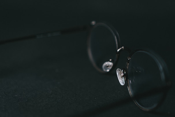 A pair of round, metal-rimmed eyeglasses lies on a dark surface with soft lighting casting subtle reflections on the lenses, creating a minimalist and sophisticated composition.