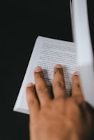 A close-up view of a person's hand gently holding the page of an open book. The background is dark, making the page and hand stand out. The text on the page is partially readable, suggesting an engaging reading experience.