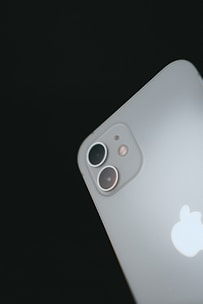 A close-up view of a sleek smartphone focusing on its dual camera lenses at the back. The Apple logo is prominently displayed below the cameras, and the surface is smooth and metallic.