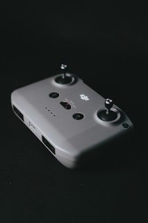 A gray drone remote controller with dual joysticks and several buttons is placed against a dark background. The design is sleek and minimalist, featuring a power button and indicator lights.