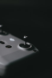 A close-up view of a joystick on a gaming or drone controller, showcasing a sleek and minimalist design. The surface is smooth with a few visible buttons, including a power button. The dark background emphasizes the controller's contours.