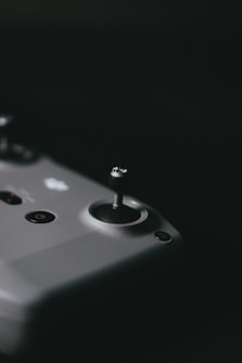 A close-up view of a joystick on a gaming or drone controller, showcasing a sleek and minimalist design. The surface is smooth with a few visible buttons, including a power button. The dark background emphasizes the controller's contours.