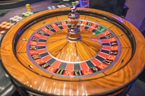 A roulette wheel with alternating red and black numbered pockets on a wooden platform. The wheel contains a small white ball resting in one of the pockets, indicating gameplay. Surrounding the wheel are stacks of colorful poker chips, suggesting a casino environment.