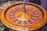 A roulette wheel with alternating red and black numbered pockets on a wooden platform. The wheel contains a small white ball resting in one of the pockets, indicating gameplay. Surrounding the wheel are stacks of colorful poker chips, suggesting a casino environment.