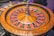 A roulette wheel with alternating red and black numbered pockets on a wooden platform. The wheel contains a small white ball resting in one of the pockets, indicating gameplay. Surrounding the wheel are stacks of colorful poker chips, suggesting a casino environment.