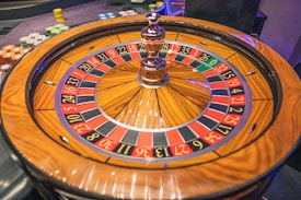 A roulette wheel with alternating red and black numbered pockets on a wooden platform. The wheel contains a small white ball resting in one of the pockets, indicating gameplay. Surrounding the wheel are stacks of colorful poker chips, suggesting a casino environment.
