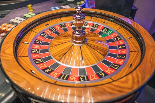 A roulette wheel with alternating red and black numbered pockets on a wooden platform. The wheel contains a small white ball resting in one of the pockets, indicating gameplay. Surrounding the wheel are stacks of colorful poker chips, suggesting a casino environment.
