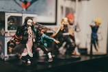 A collection of detailed anime figurines displayed on a shelf. The figures represent various characters in dynamic poses, surrounded by boxes with similar designs. The focus is on a female character with long dark hair and a pink patterned outfit. Background figures include characters with vibrant costumes and distinctive hairstyles.