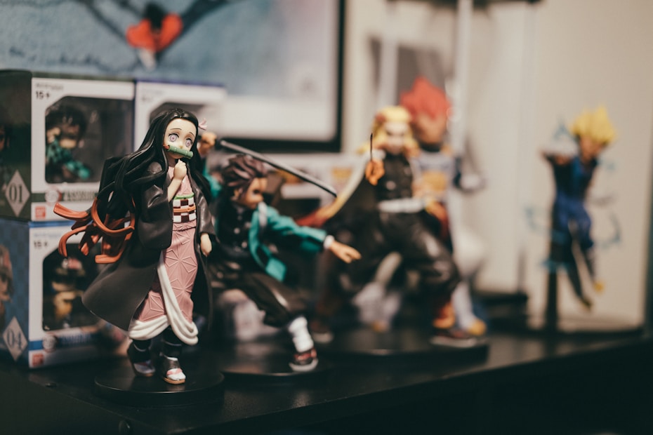 A collection of detailed anime figurines displayed on a shelf. The figures represent various characters in dynamic poses, surrounded by boxes with similar designs. The focus is on a female character with long dark hair and a pink patterned outfit. Background figures include characters with vibrant costumes and distinctive hairstyles.