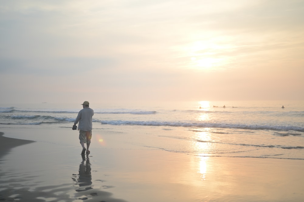 homem na camisa branca e calças pretas andando na praia durante o pôr do sol