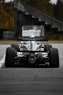 A high-performance race car with the Renault Sport logo displayed prominently on the back. The car is stationary on a track, viewed from behind, with large, smooth treaded tires. In the background, there is a blurred structure and some greenery.