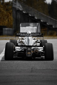A high-performance race car with the Renault Sport logo displayed prominently on the back. The car is stationary on a track, viewed from behind, with large, smooth treaded tires. In the background, there is a blurred structure and some greenery.
