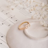 A delicate gold ring with small diamond accents rests on a textured white ceramic dish, surrounded by fine sheets of music notes. Sprigs of small white flowers add a soft, romantic touch to the composition.