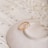 A delicate gold ring with small diamond accents rests on a textured white ceramic dish, surrounded by fine sheets of music notes. Sprigs of small white flowers add a soft, romantic touch to the composition.