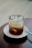 A glass filled with iced coffee or a cold beverage sits atop a white saucer on a wooden table. The drink contains visible ice cubes and varying shades of brown liquid.
