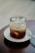 A glass filled with iced coffee or a cold beverage sits atop a white saucer on a wooden table. The drink contains visible ice cubes and varying shades of brown liquid.