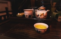A ceramic teapot and a matching cup are placed on a dark wooden surface. The teapot features a design with roosters, and steam is gently rising from the tea in the cup. The setting has a warm, cozy ambiance with dim lighting.