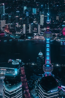 A nighttime cityscape featuring a futuristic skyline with bright neon lights illuminating the buildings. The large, distinctive tower with a spherical structure stands prominently in the foreground, casting a soft glow on the surrounding high-rise architecture. The city appears vibrant and bustling, reflected in the calm water below.