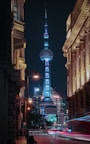 A cityscape at night featuring the iconic Oriental Pearl Tower, surrounded by illuminated skyscrapers and historic buildings. Streets are lively with blurred motion from passing vehicles and the warm glow of street lamps contrasting with the cool blue and white lights of the tower.