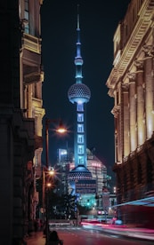 A cityscape at night featuring the iconic Oriental Pearl Tower, surrounded by illuminated skyscrapers and historic buildings. Streets are lively with blurred motion from passing vehicles and the warm glow of street lamps contrasting with the cool blue and white lights of the tower.