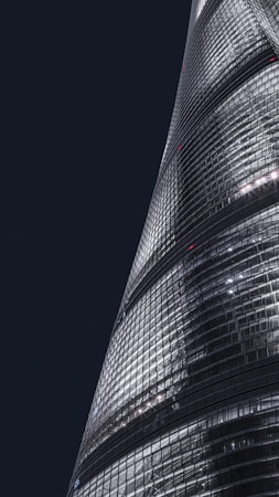 A sleek, modern skyscraper with a curved, glass facade is illuminated against a dark night sky. The building's surface reflects bright artificial lights, creating a striking contrast with the surrounding darkness.