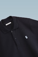This is a close-up image of a black polo shirt with a collar and two buttons. There is a small embroidered design on the chest, consisting of two overlapping shapes in light blue. The label inside the collar reads 'SCAMBIO'. The background is a simple gradient from pale blue to white.