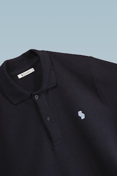 This is a close-up image of a black polo shirt with a collar and two buttons. There is a small embroidered design on the chest, consisting of two overlapping shapes in light blue. The label inside the collar reads 'SCAMBIO'. The background is a simple gradient from pale blue to white.