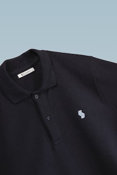 This is a close-up image of a black polo shirt with a collar and two buttons. There is a small embroidered design on the chest, consisting of two overlapping shapes in light blue. The label inside the collar reads 'SCAMBIO'. The background is a simple gradient from pale blue to white.