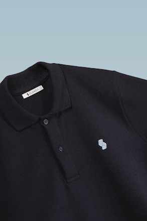 This is a close-up image of a black polo shirt with a collar and two buttons. There is a small embroidered design on the chest, consisting of two overlapping shapes in light blue. The label inside the collar reads 'SCAMBIO'. The background is a simple gradient from pale blue to white.