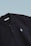 This is a close-up image of a black polo shirt with a collar and two buttons. There is a small embroidered design on the chest, consisting of two overlapping shapes in light blue. The label inside the collar reads 'SCAMBIO'. The background is a simple gradient from pale blue to white.