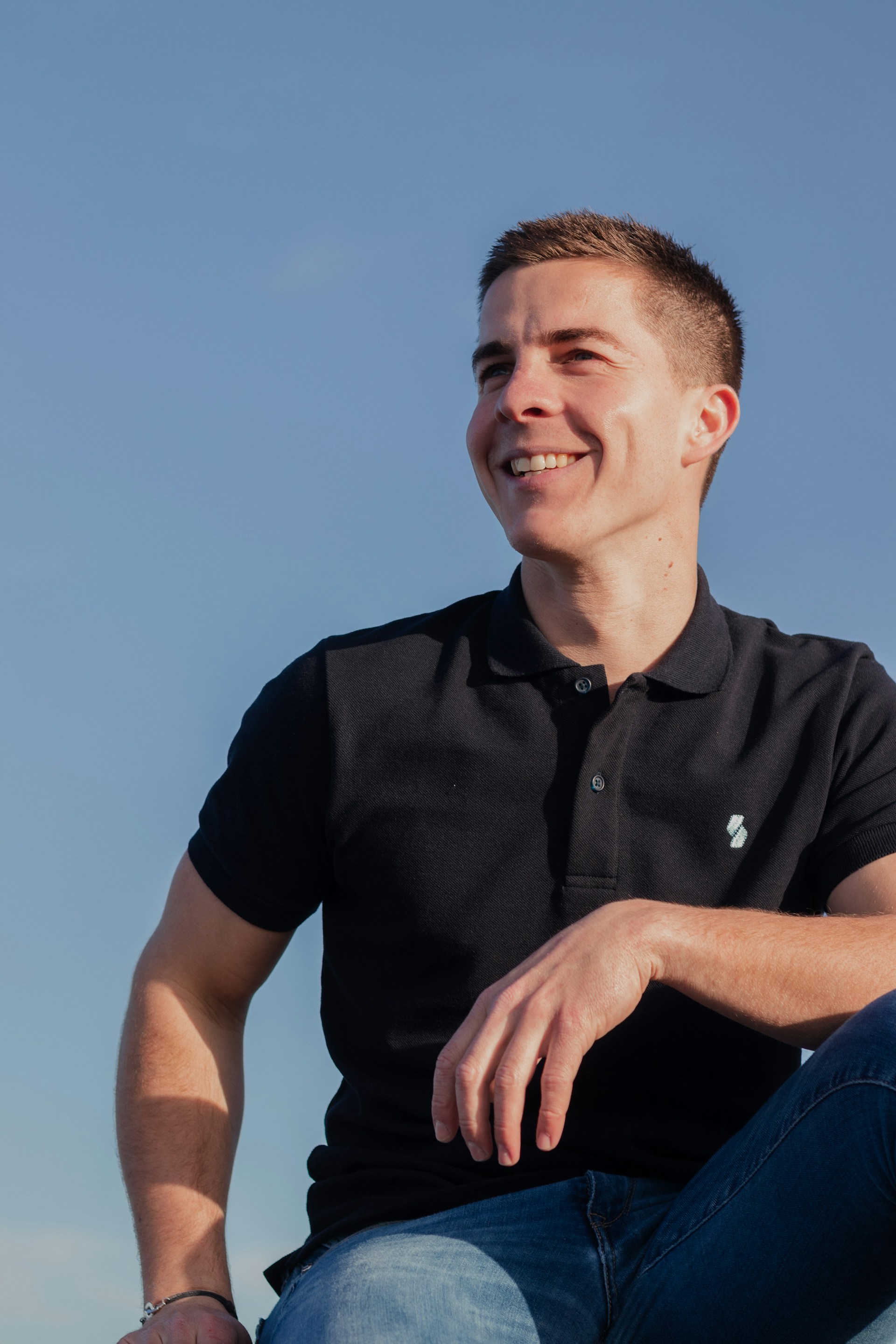 A cheerful model wearing a sky-blue polo shirt, standing casually against a sunlit brick wall.