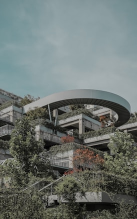 A modern, multi-level building structure is integrated with lush greenery. The architecture includes multiple terraces filled with various types of vegetation, including shrubs and small trees. An architectural feature resembling a circular overhang is present at the top, adding a unique element to the design. The sky is a soft, pale blue, suggesting a calm atmosphere.