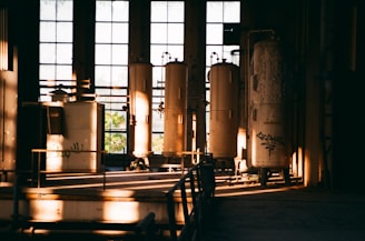 A dimly lit industrial space with large cylindrical tanks and machinery inside an old building. The sunlight streaming through tall, dusty windows casts long shadows, creating a dramatic play of light and shadow. Some graffiti can be seen on one of the tanks, adding a touch of urban decay.